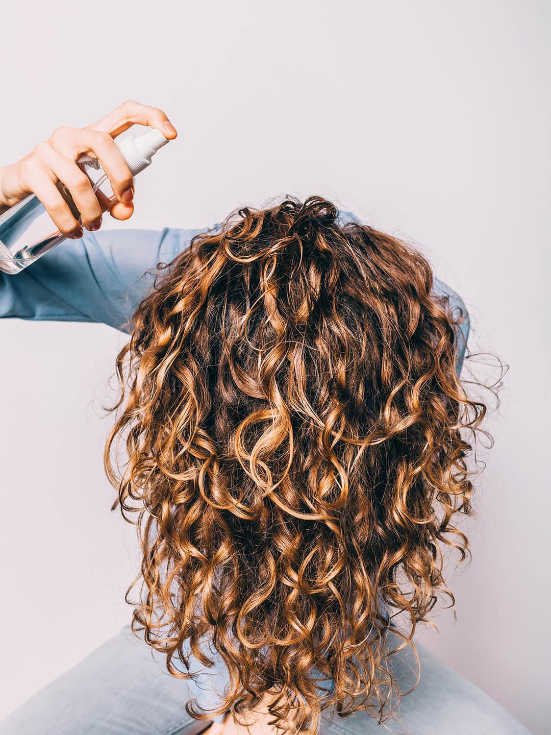 Woman spraying hair care product on her long curly hair over her head Desktop