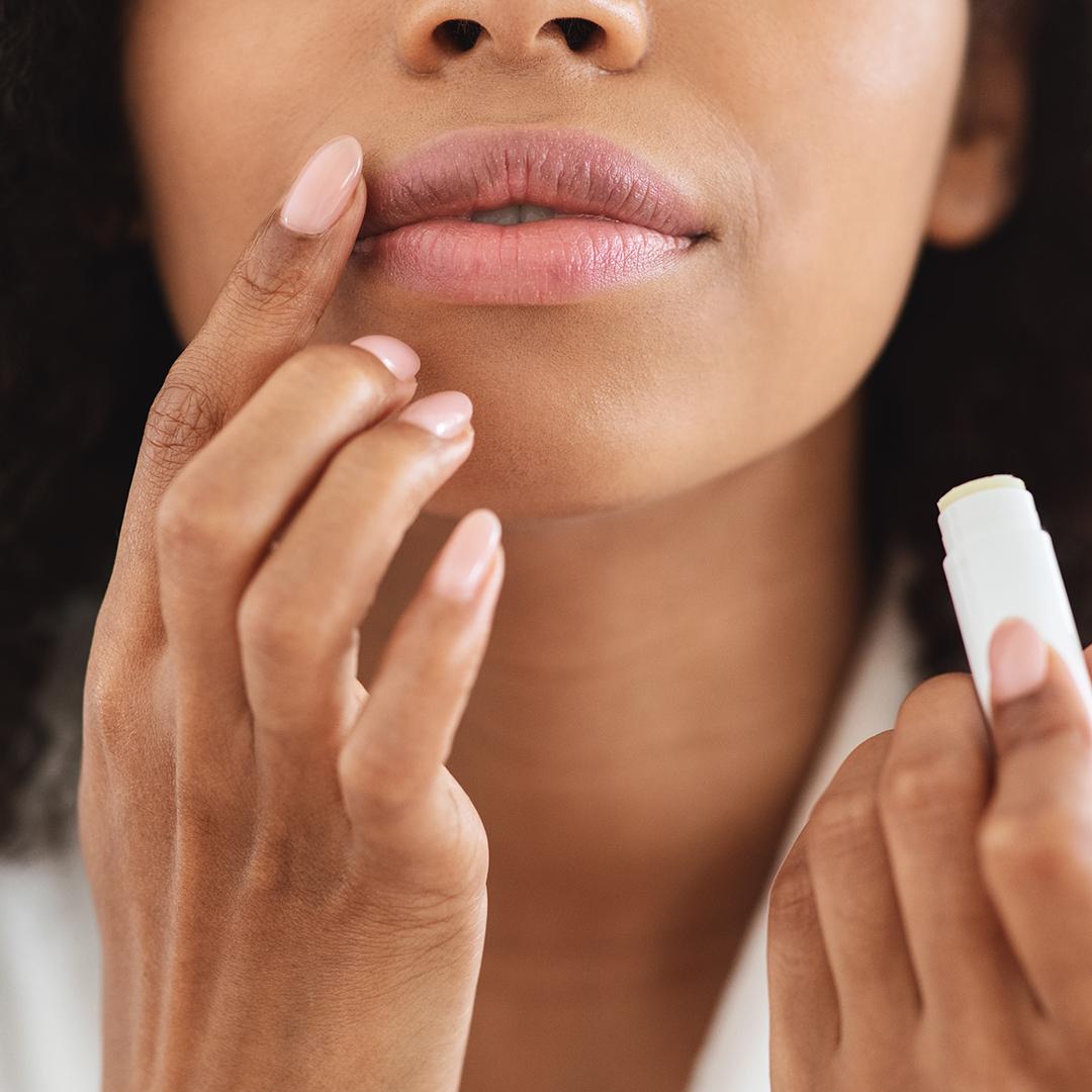 An image of a woman focused on touching the side part of her lips while holding a white lip chapstick Mobile