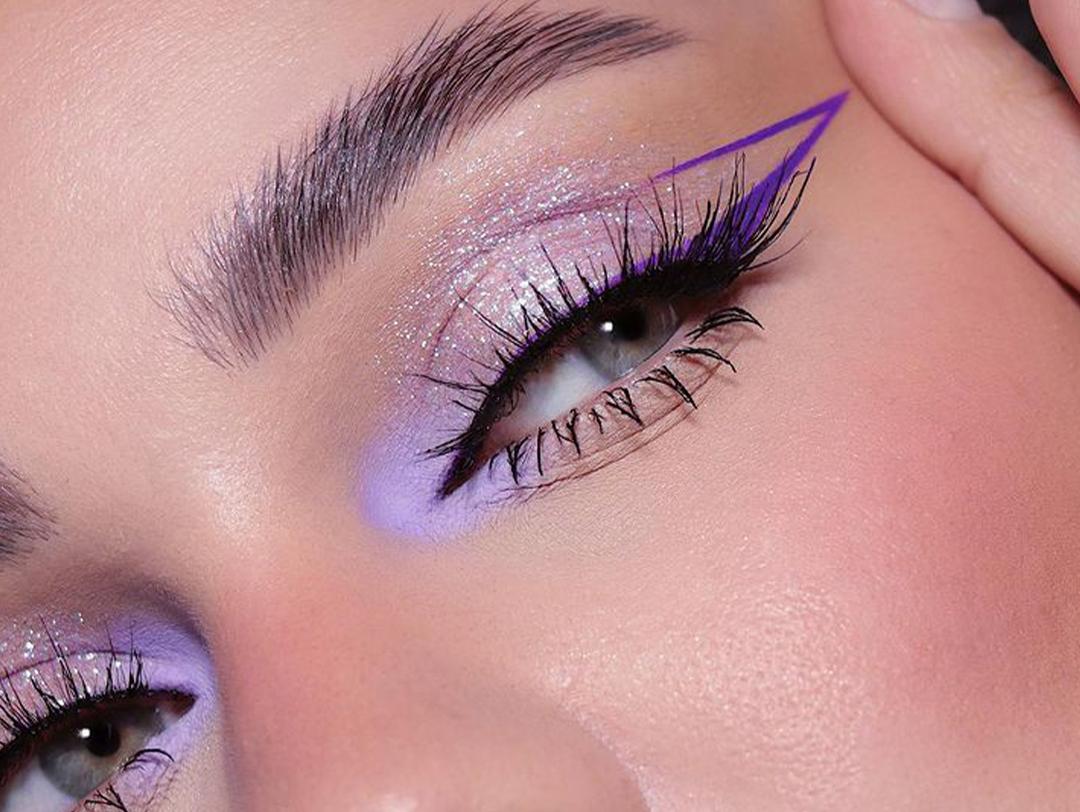 A close-up photo of a woman wearing a purple- winged eyeliner paired with glossy nude lip color. Desktop