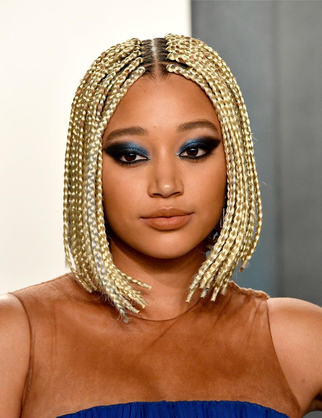 Amandla Stenberg looking glam in a blue dress with sheer neckline, shimmery dark blue smokey eye look, and blonde braided bob hairstyle Mobile