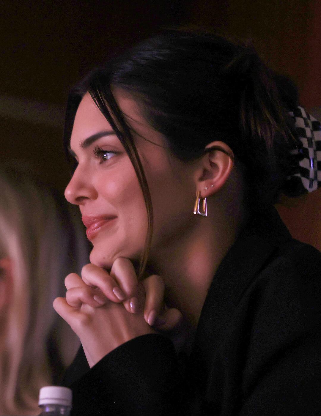 A photo of Kendall Jenner smiling with her hair styled in a claw clip updo wearing a black trench coat and gold earrings Desktop