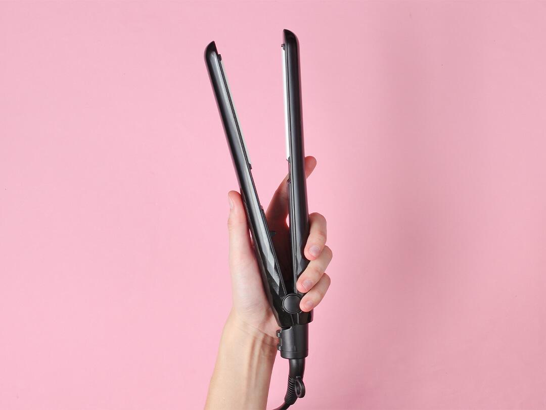 Image of a hand holding up a hair flat iron against a pink background Desktop