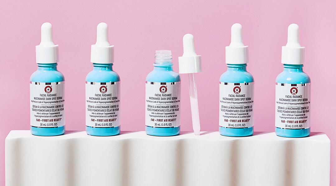 Row of FIRST AID BEAUTY Facial Radiance Niacinamide Dark Spot Serum on white block against pink background Desktop