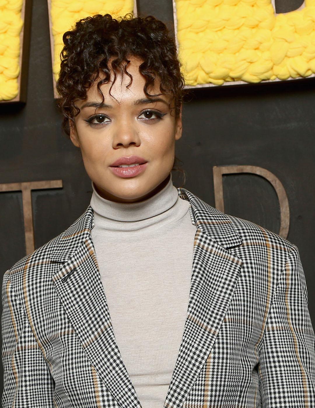 A photo of Tessa Thompson in her ivory-colored inner turtleneck topped with a stripe coat that accentuates her dark curly fringe hair on top of her face Desktop