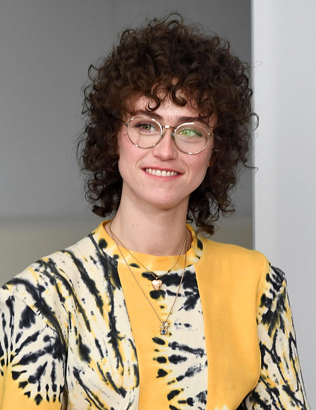 A photo of Ella Emhoff in her black curly mullet wearing a round-wired eyeglasses, yellow tie-dye T-shirt, and black pants Mobile