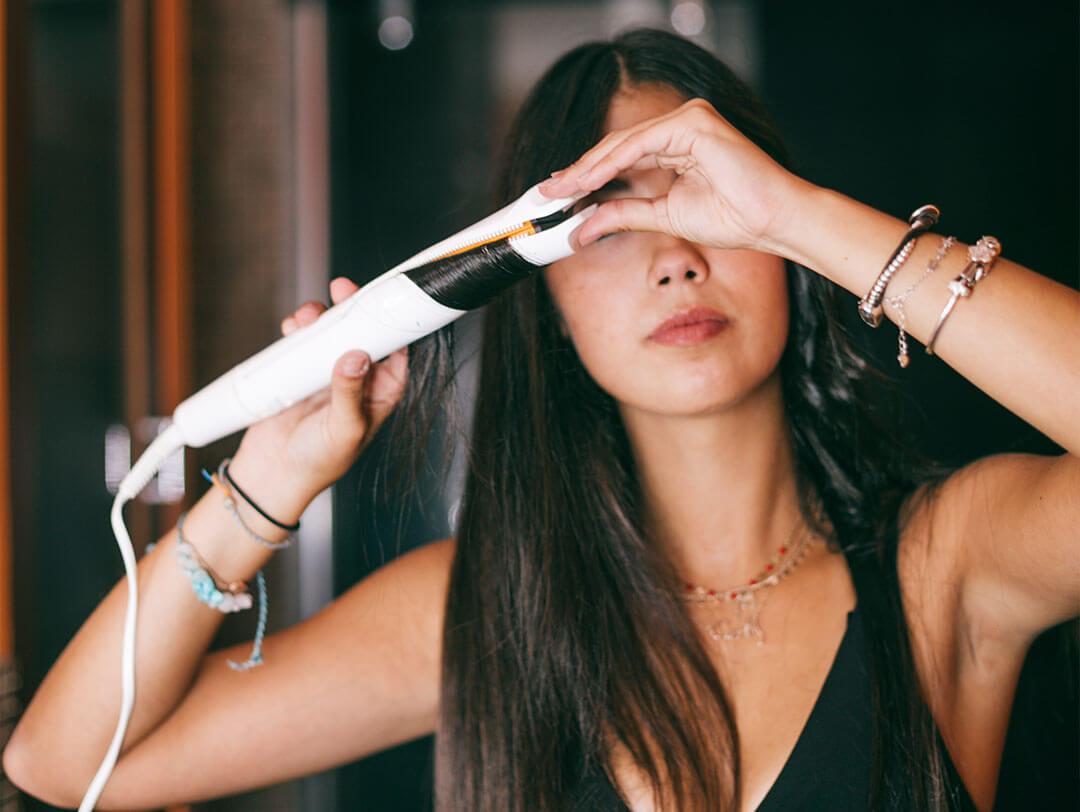 Brunette young woman styling her long hair with curling iron or straightener, exuding elegance in a stunning black dress Desktop
