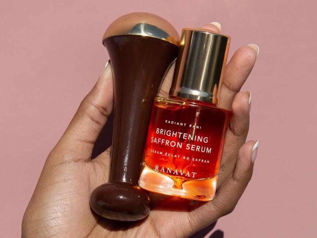 A photo of a woman's hand holding Kansa Wand and Brightening Saffron Serum on a rose gold background Desktop