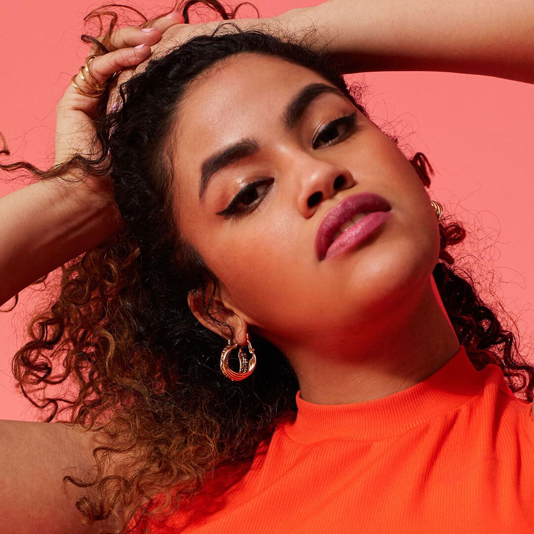 Close-up image of a model with thick, curly hair rocking a natural makeup look wearing an orange top on a red background Desktop