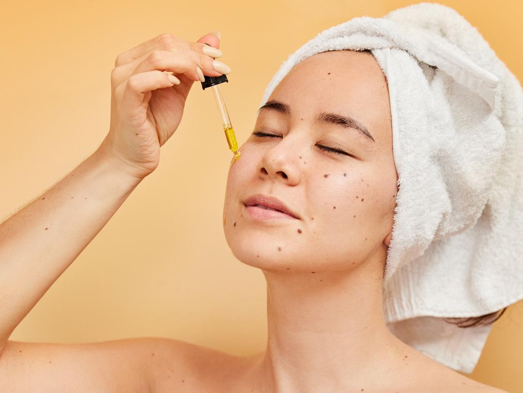 A photo of a woman applying serum on her cheek with a white towel on her head on a yellow background Desktop