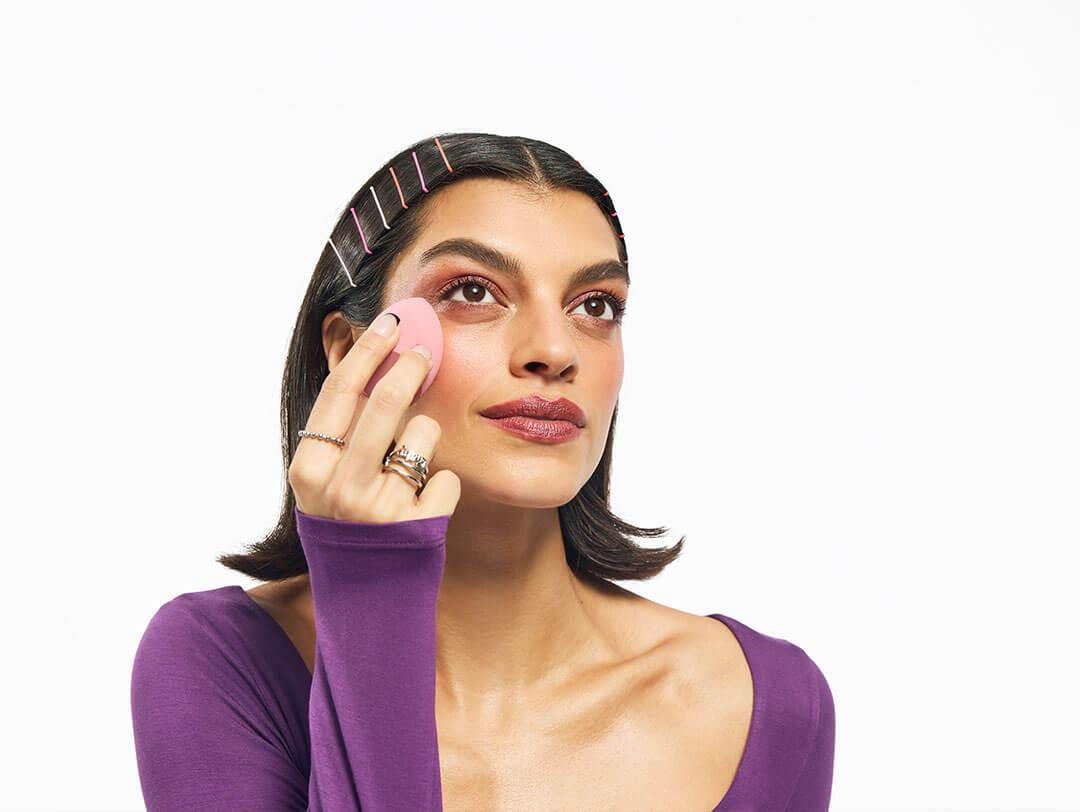 An image of a woman sporting shoulder-length hair fastened with vibrant bobby pins, clad in a long-sleeve purple top, is holding a beauty sponge Desktop