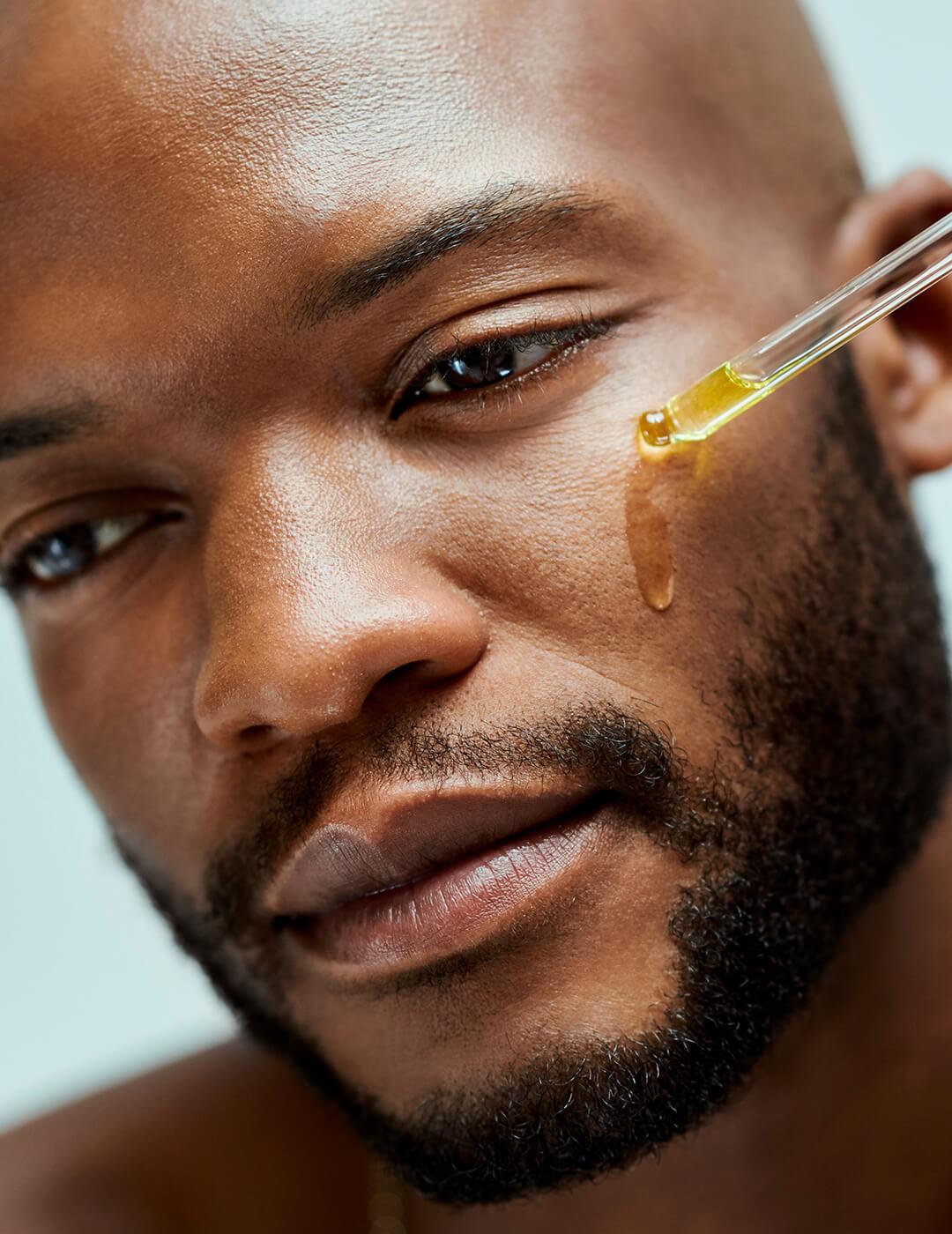 African American applying serum on his face using a dropper. Attractive male model with a beard inside of a room with an isolated studio background. He is wearing a necklace Desktop