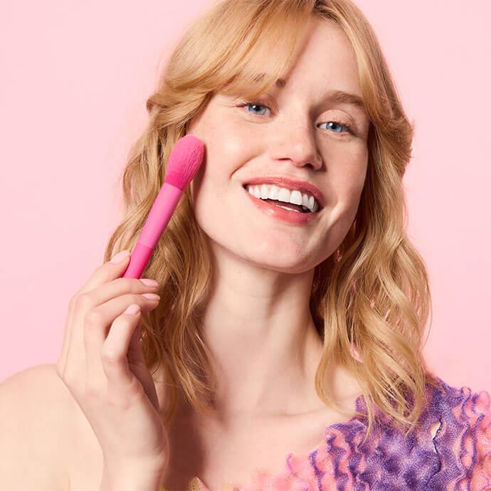 A woman with wavy blonde hair applies blush to her cheek using a bright pink makeup brush against a soft pink background