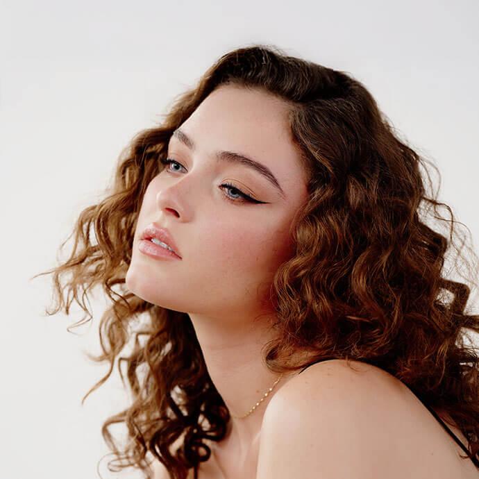 A portrait of a woman with curly brown hair and natural makeup—winged eyeliner, rosy cheeks, glossy nude lips, subtle highlighter, and neutral eyeshadow—gazing upward with a serene expression